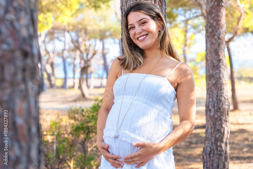 Portrait of smiling pregnant woman in white summer dress, standing on beach near palm tree and looking at camera.natural outdoor setting and happiness reflect calm maternity lifestyle moment.