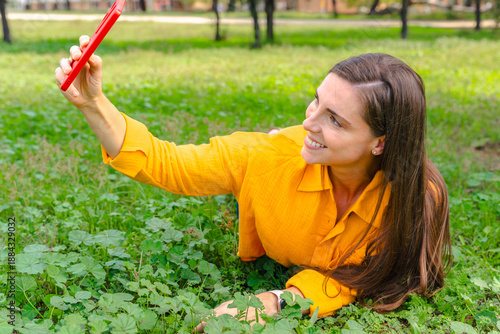 Happy woman wearing yellow shirt lying on green grass outdoors taking selfie with smartphone, concept of leisure, social media, connection, positivity, nature, lifestyle, relaxation.