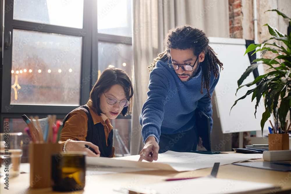 Obraz premium Young Caucasian woman with disability sitting at desk working with blueprints while adult Black man standing beside her pointing at paper, both collaborating on architectural project in modern office