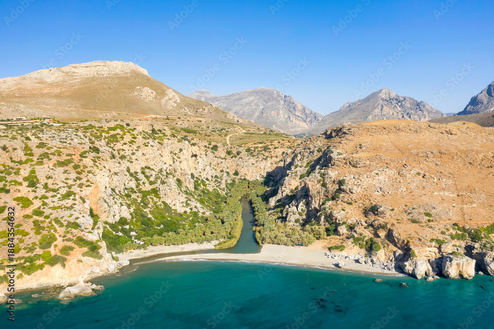 Naklejka premium Aerial perspective of a sandy beach where a winding river meets the turquoise sea, framed by steep rocky cliffs and sparse Mediterranean vegetation near Preveli, Crete. Bright sunlight and clear skies