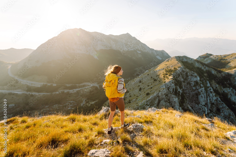 Obraz premium A young woman with hiking backpack stands on mountain trail on sunny day. Female hiker with a yellow backpack enjoys mountain scenery at sunset. Concepts of adventure, freedom. Active lifestyle.