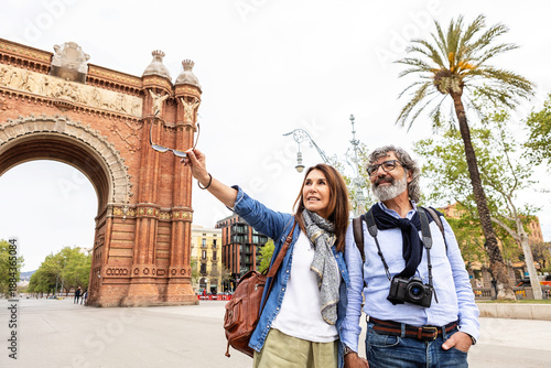 Senior adult married couple happily exploring barcelona, with the iconic brick archway of the city's landmark in the background and a palm tree framing the scene