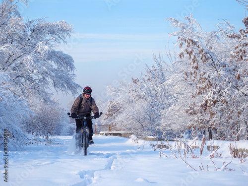 Male cyclist riding a mountain bike through a snow-covered path surrounded by frosty trees under a clear blue sky in a winter landscape