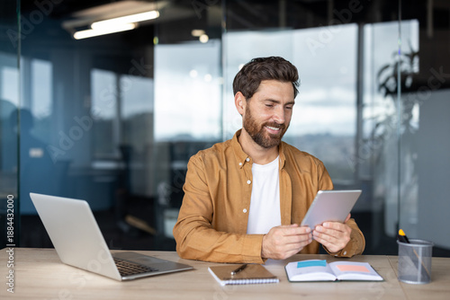 Smiling young man with a beard actively engaging with a digital tablet at a desk, surrounded by a laptop, notebooks, and office supplies in a contemporary workspace