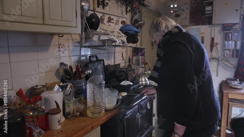 A woman prepares a meal in her cluttered kitchen. She moves around with utensils, checking pots and getting ingredients ready for cooking. The setting shows signs of daily life.