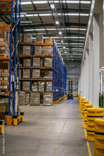 Wide warehouse aisle with tall storage racks stacked with pallets of various cardboard boxes, showcasing organised industrial logistics and inventory management.