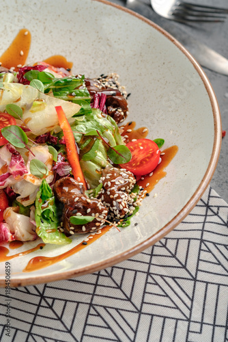 Asian-inspired salad with glazed teriyaki beef chunks in sesame seeds, fresh mixed greens, cherry tomatoes, carrot strips and soy dressing in rustic white bowl. Vibrant fusion gourmet dish