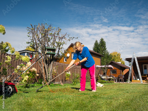 Canvas Print Woman raking cut grass in a lush green backyard garden near a wooden house