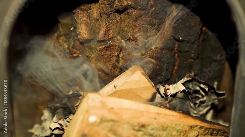 Burning paper and cardboard with smoke inside metal container. Close-up macro shot of smoldering paper waste and ash.