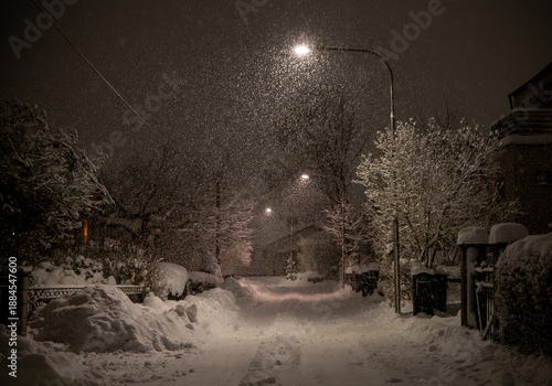 A snowy residential street during heavy nighttime snowfall, illuminated by streetlights and lined with trees and houses covered in fresh winter snow.