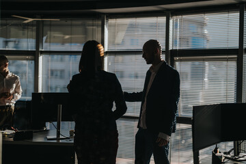 Two professionals in an office scene converse near desks and computer monitors. The setting features large windows with blinds, creating silhouettes and a professional atmosphere.