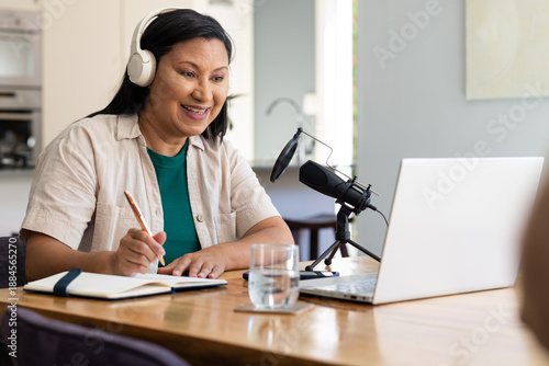 Woman wearing white headset speaking into microphone with pop filter, taking notes at dining table