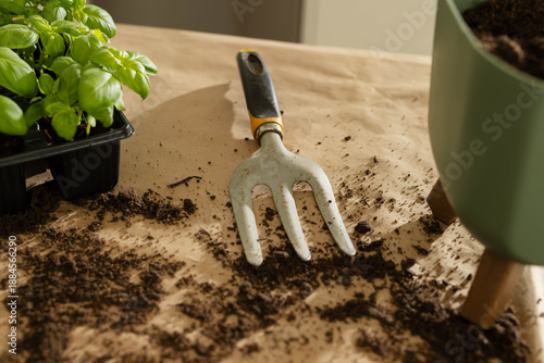 Hand cultivator is resting on kraft paper, soil clinging, seed tray and green pot near window