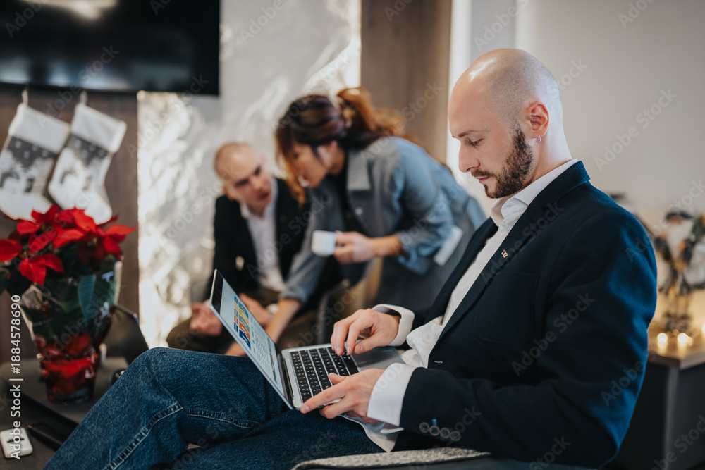 Fototapeta premium A man in a blazer works on his laptop, while coworkers chat in the background. The scene captures a modern office holiday atmosphere with warm lighting and festive decor.