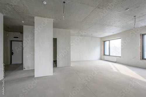 Spacious room with white box pre-finish: concrete ceiling floor, plastered walls, partition columns, large window, radiator, exposed wiring, minimal fixtures, ready for design.