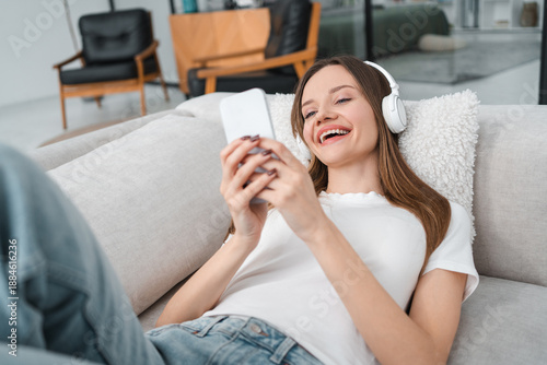Girl, smartphone and smile with mobile app for listening to music on couch at apartment. Woman, phone and headphones on sofa, happy and texting in home with streaming, audio and relax in lounge.