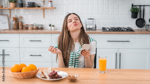 Young happy woman sitting at the table in the kitchen, eating sandwich with fruits and drinking juice, having breakfast, enjoying mindful morning routine and lifestyle. Home, wellness, healthy habits.
