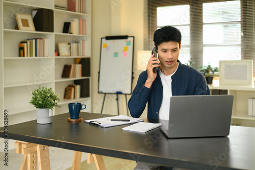 Young Asian businessman talking on the phone while working on a laptop in a home office, representing online negotiation and remote business