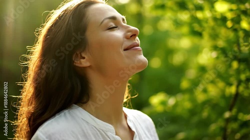 Woman breathing fresh air in forest - A smiling woman with her eyes closed takes a deep breath in a lush green forest with sunlight streaming through the trees.