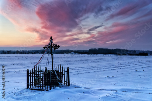 Winter rural landscape at sunset. A roadside shrine in the foreground. Dramatic purple clouds in the sky. A field covered with snow.  Podlachia, Poland, Europe.