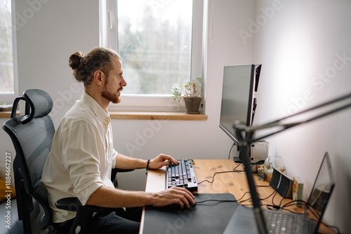 Man works on computer at home office with natural light, focused on tasks during daytime, surrounded by plants and modern furniture