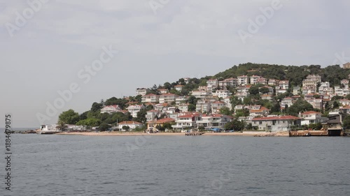 A scenic view of a coastal village on the Princes Islands in Istanbul, Turkey, with houses nestled among green trees and overlooking calm waters.