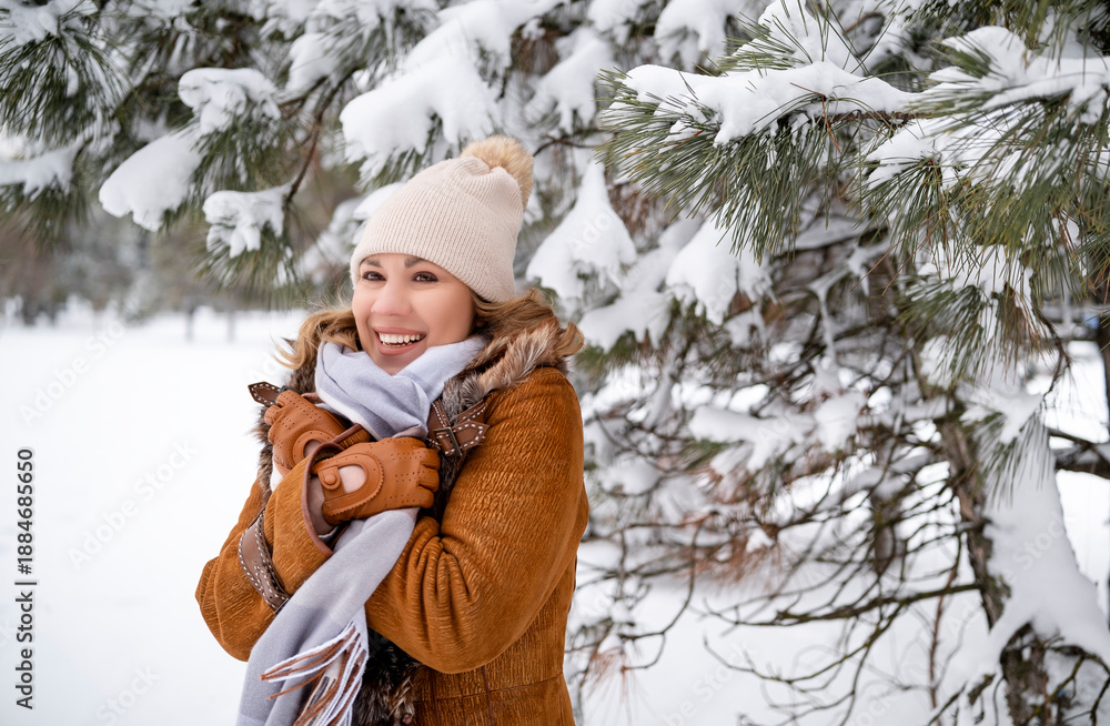 Obraz premium Smiling middle aged woman in warm brown sheepskin coat and white hat in winter forest. Beautiful female enjoys snowy nature. Christmas, travel, weekend, lifestyle concept