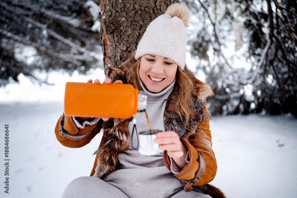 Fototapeta premium Smiling woman pours hot tea from orange thermos in winter forest. Middle aged female sits near tree on snow. Picnic, relax, nature, weekend, wellness concept