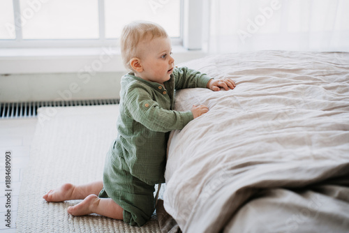 фотографія A curious baby kneels near a bed, exploring its surroundings with wide-eyed wonderment in a softly lit, cozy room