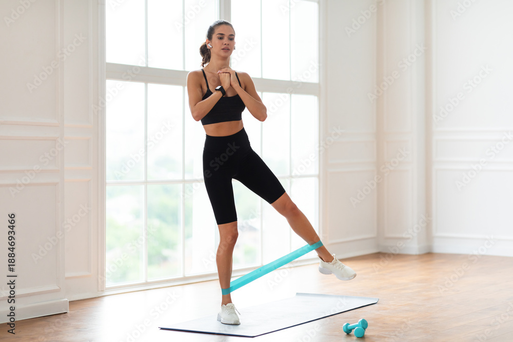 Fototapeta premium Full And Lower Body Workout. Focused Sporty Woman Exercising With Elastic Rubber Band Lifting And Kicking Leg Aside Standing On Yoga Mat, Listening To Music In Wireless Earpods. Bodyweight Training