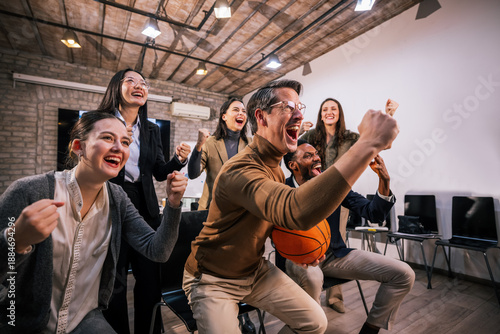 Business team watching a basketball game together at the office, cheering with excitement while one coworker holds a basketball, representing teamwork, relaxation, and positive work atmosphere.