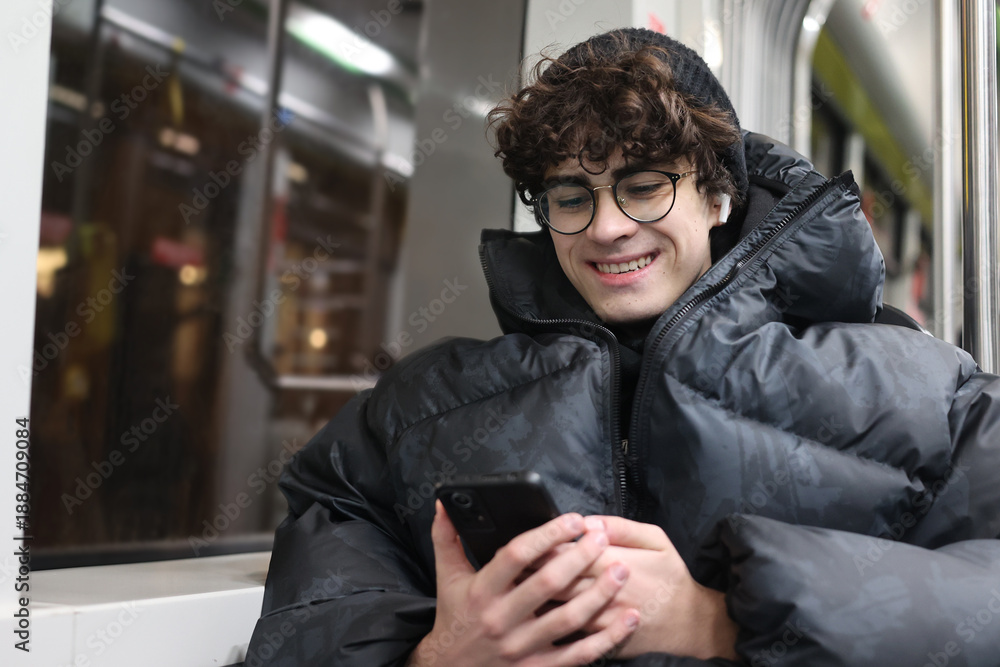 Obraz premium Smiling adolescent using mobile phone while commuting in the city. Modern youth lifestyle, daily commute, technology use, urban life and mobility concept.