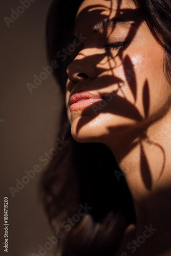Woman with long dark hair, illuminated by soft light, has shadows cast on her face from nearby plants, creating a serene and artistic atmosphere in a minimalist setting