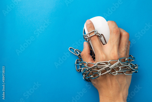 A hand holding a computer mouse is tied with metal chains on a blue background. Concept for internet addiction, cybercrime, digital slavery, and online security constraints with copy space.