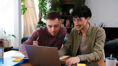 Two young men collaborating, using a laptop and writing notes in their living room