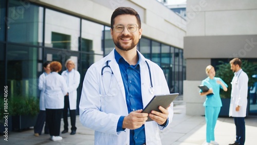 Wallpaper Mural Confident Caucasian male doctor standing outside hospital. Medical specialist holding tablet. Background colleagues discussing cases. Outdoor clinic area hosting professional team. Torontodigital.ca
