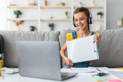 Принти на полотні Smiling girl having video call with teacher, holding flipchart with paper showing done task result using laptop