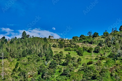 Moon rises over lush green mountains with a scenic Ecuadorian mirador, showcasing vibrant vegetation and a clear blue sky during twilight hours with copy space