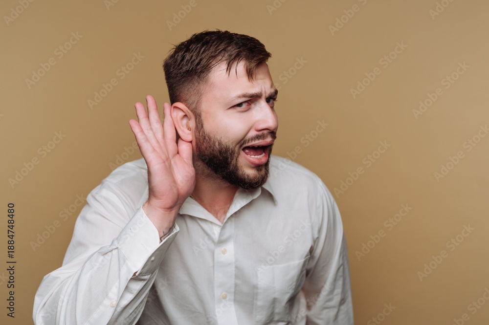 Fototapeta premium Man in white shirt cupping ear, listening intently, beige background.