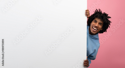 Schilderij op canvas Funky black teen guy peeking out from behind blank white paper banner with mockup for design, pink studio background