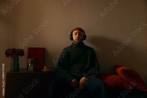 Photos A man sits in a dimly lit room, wearing a beanie and headphones with his eyes closed, enjoying a peaceful moment of music or meditation in a cozy, atmospheric interior