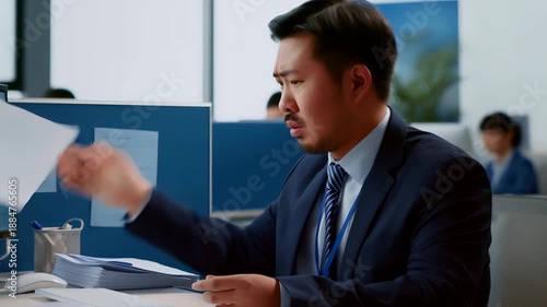 Asian businessman. Dressed in a suit. Sitting at his desk in an open plan office. Holding and reading a document with a confused and stressed expression. Looking through data and challenging paperwork