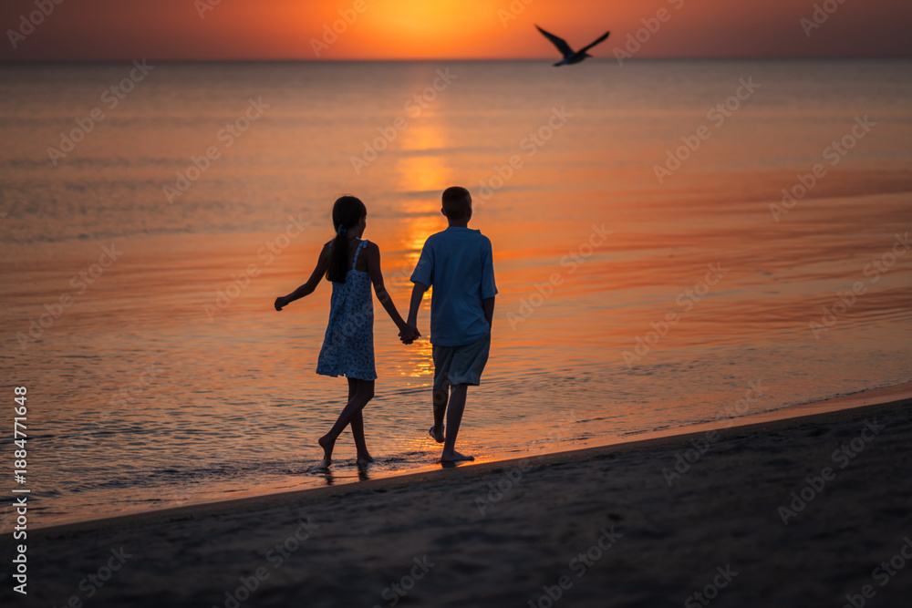 Fototapeta premium Siblings play around the beach enjoying waves and sun. Children walk along the beach at sunset holding hands. High quality photo