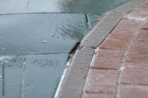 Ice-covered pavement showing a transition between gray concrete and red brick, with a thin layer of freezing rain creating a glossy surface in a residential area