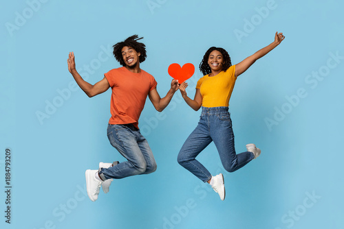Happy african american young man and woman loving couple jumping up with heart-shaped red card over blue studio background, black lovers celebrating St. Valentines Day together