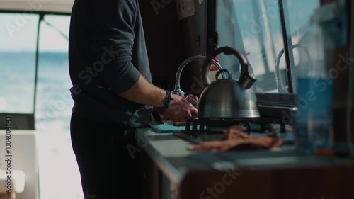 Brutal man with sleeves of black jumper rolled up washes dishes in yacht's galley next to stove with kettle on it. Close-up of his hands. Blurred background of sea bay is behind him