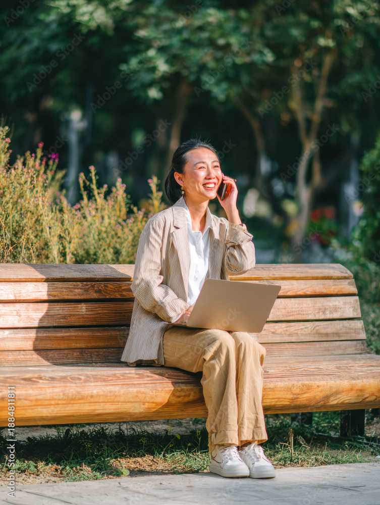 Fototapeta premium Young woman smiling and talking on the phone while working on a laptop outdoors sitting on a wooden park bench surrounded by green trees and bushes on a sunny day