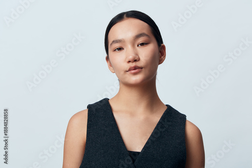 Bilde på lerret Portrait of young woman with neutral expression wearing dark sleeveless top against plain light background, looking directly at camera in studio setting