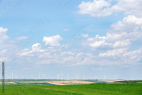 Wide panoramic view of modern wind turbines installed across agricultural fields under a bright blue sky with soft clouds. 