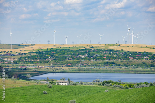 Wide landscape view of modern wind turbines installed on hills above a rural village, agricultural fields, and a calm lake.